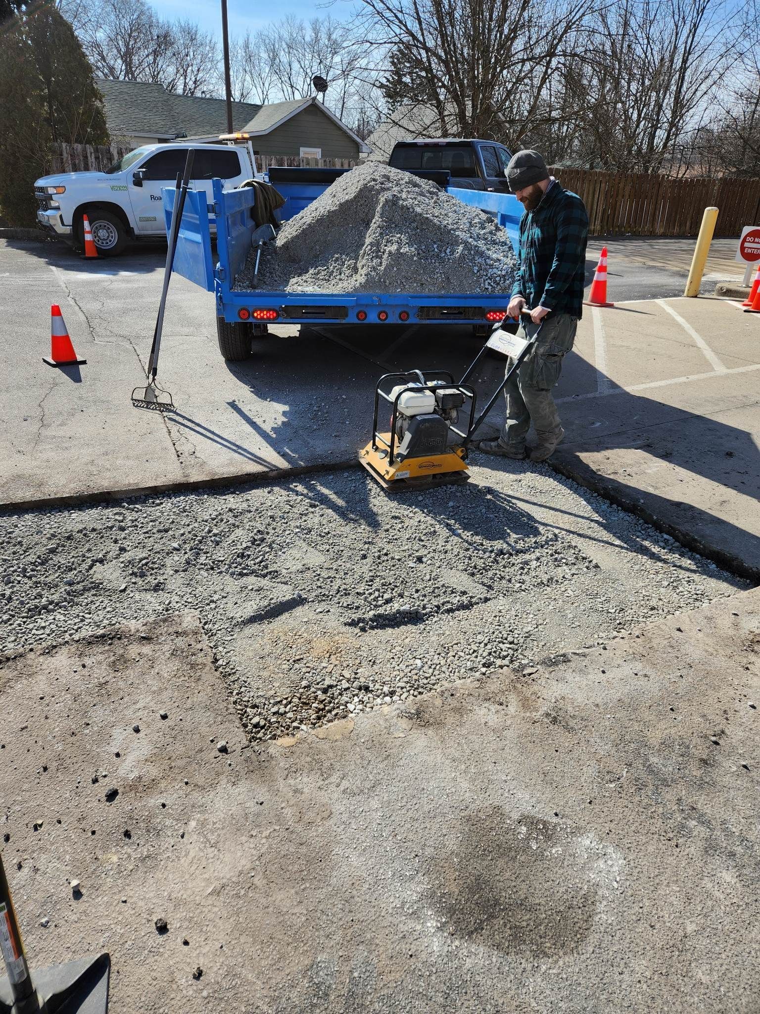 Man compacting gravel with a vibratory plate compactor to repair a parking lot. Blue truck, orange cones.