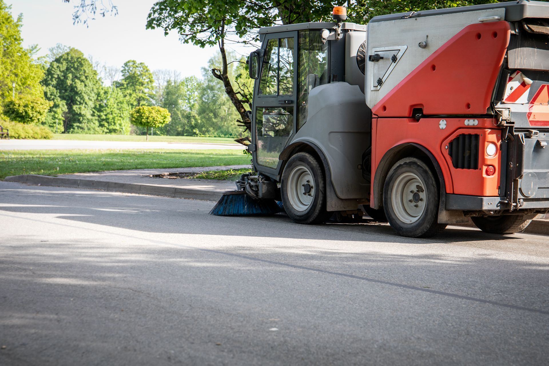 Parking lot sweeping service vehicle cleaning a paved parking area in a landscaped commercial setting.