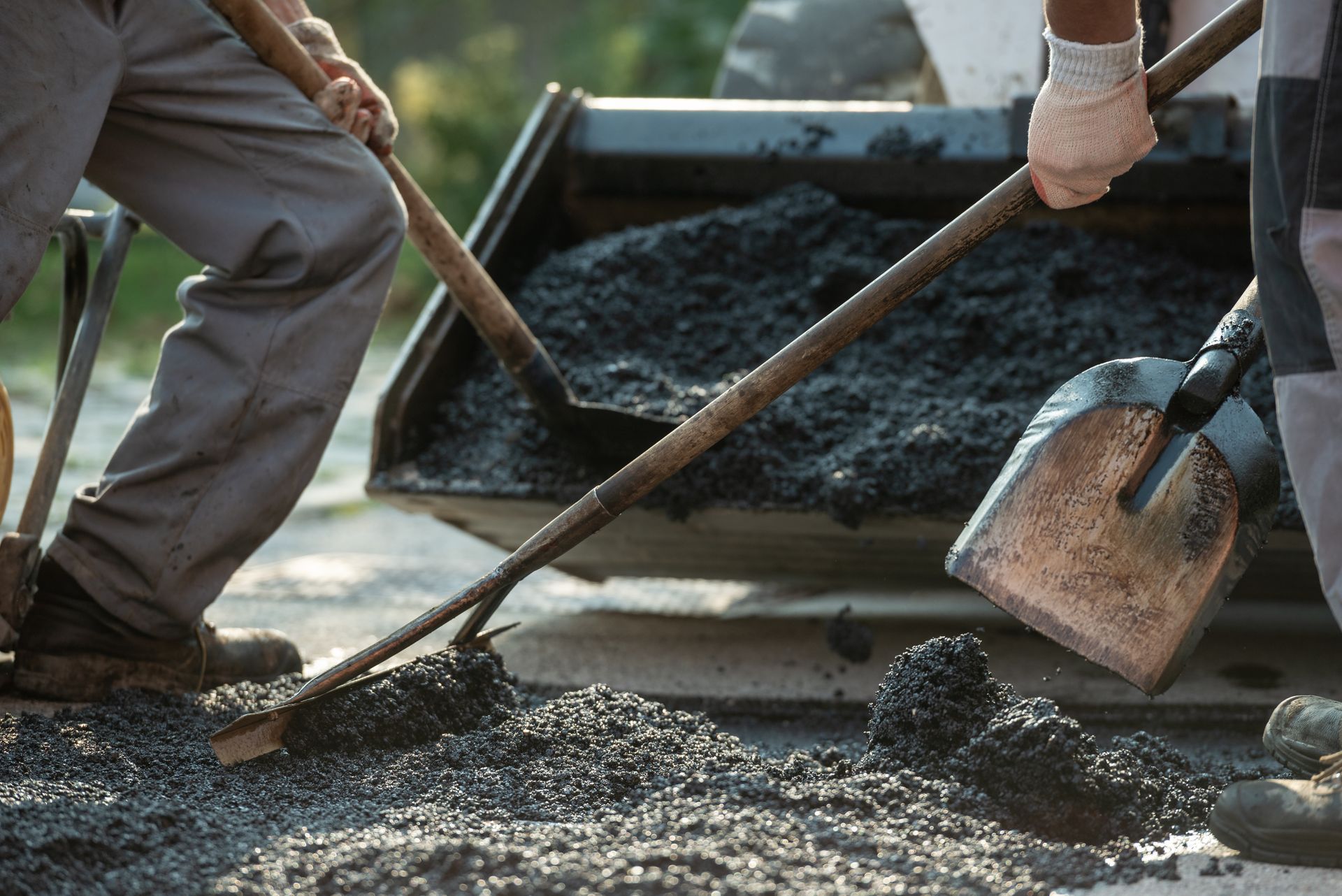 Workers spreading fresh asphalt on a road with shovels near paving equipment.