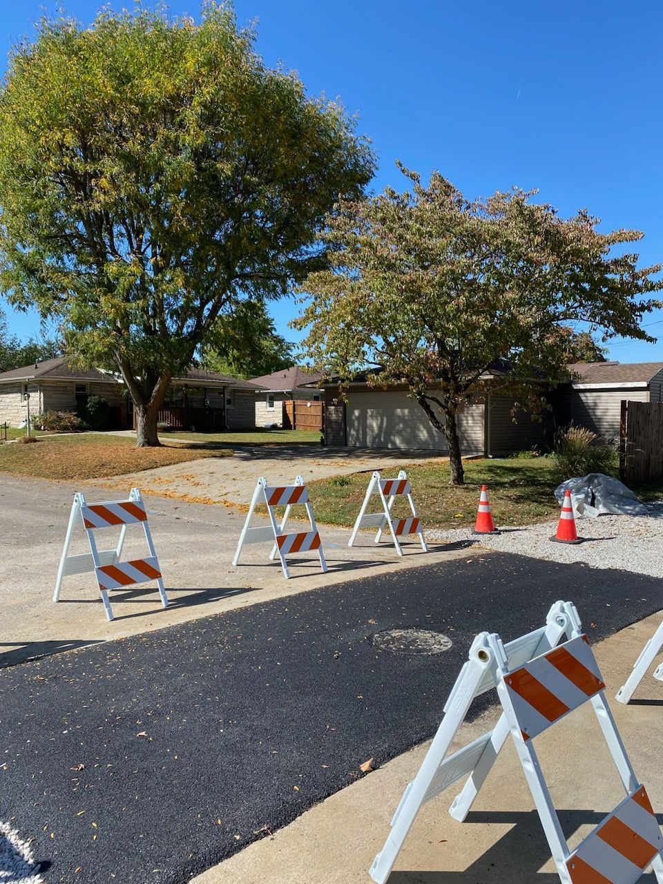 Road construction with orange and white traffic barriers blocking a newly paved surface. Houses and trees in the background.