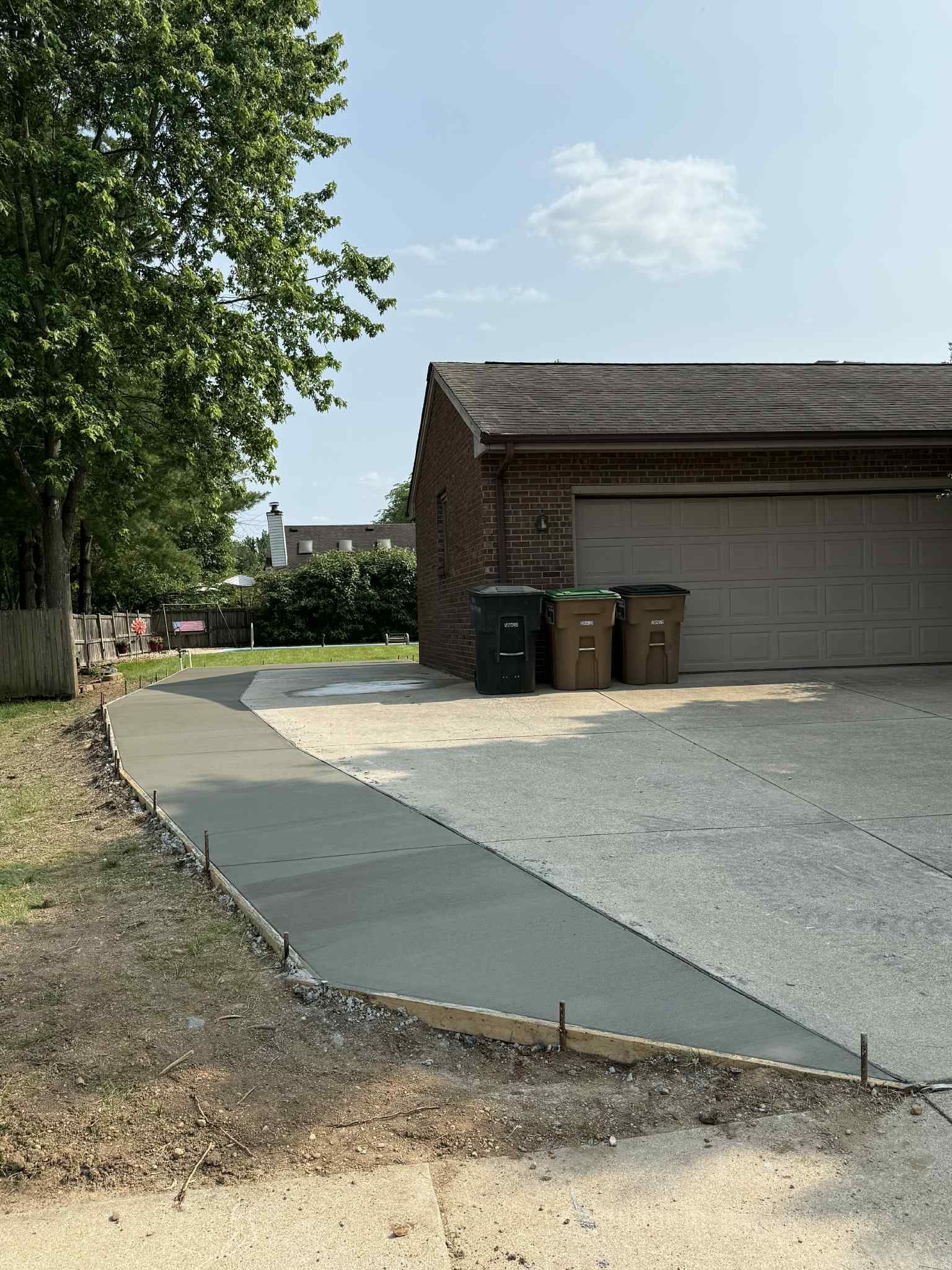 A new concrete sidewalk extends from a gravel driveway toward a brick garage and a tree on a sunny day.