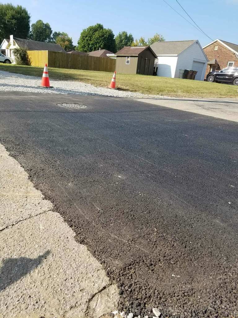 Newly paved asphalt road with orange traffic cones, houses in the background.