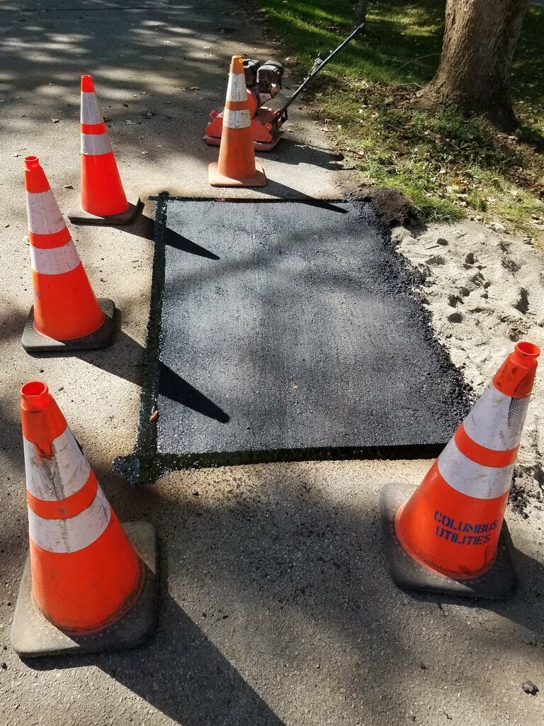 Asphalt patch on sidewalk, surrounded by orange traffic cones and a compactor.