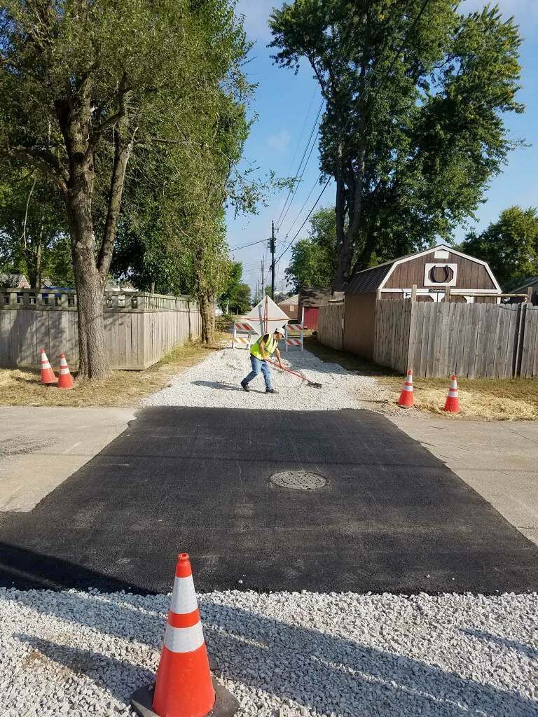 Worker paving a residential alley with gravel and asphalt; cones line the sides.