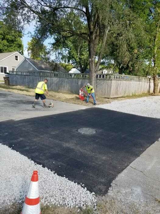 Two workers paving a street patch, using tools and wearing vests. Gray houses and a fence in the background.