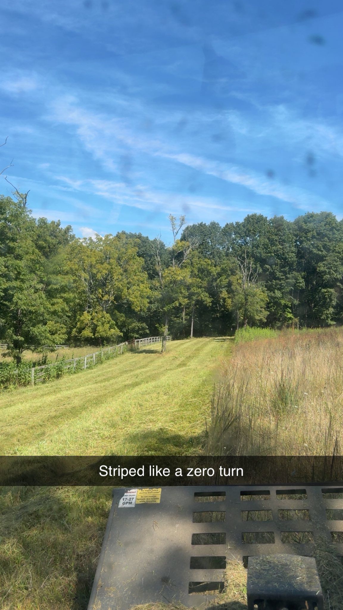 A person mowing a field with a zero-turn mower under a blue sky with trees.