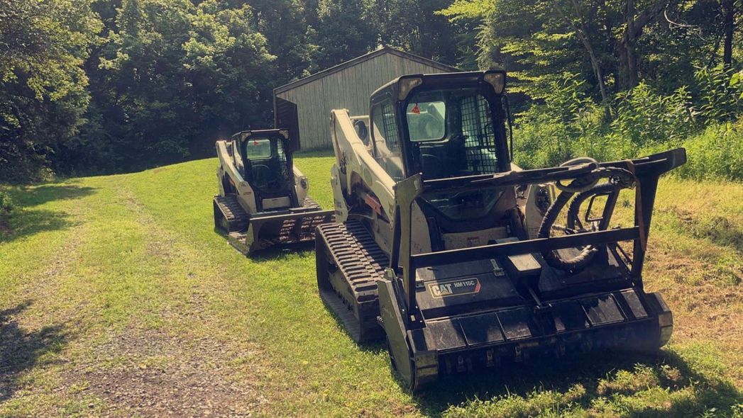 Two Bobcat skid steers on a grassy path near a small shed and trees. One has a forestry mulcher attachment.