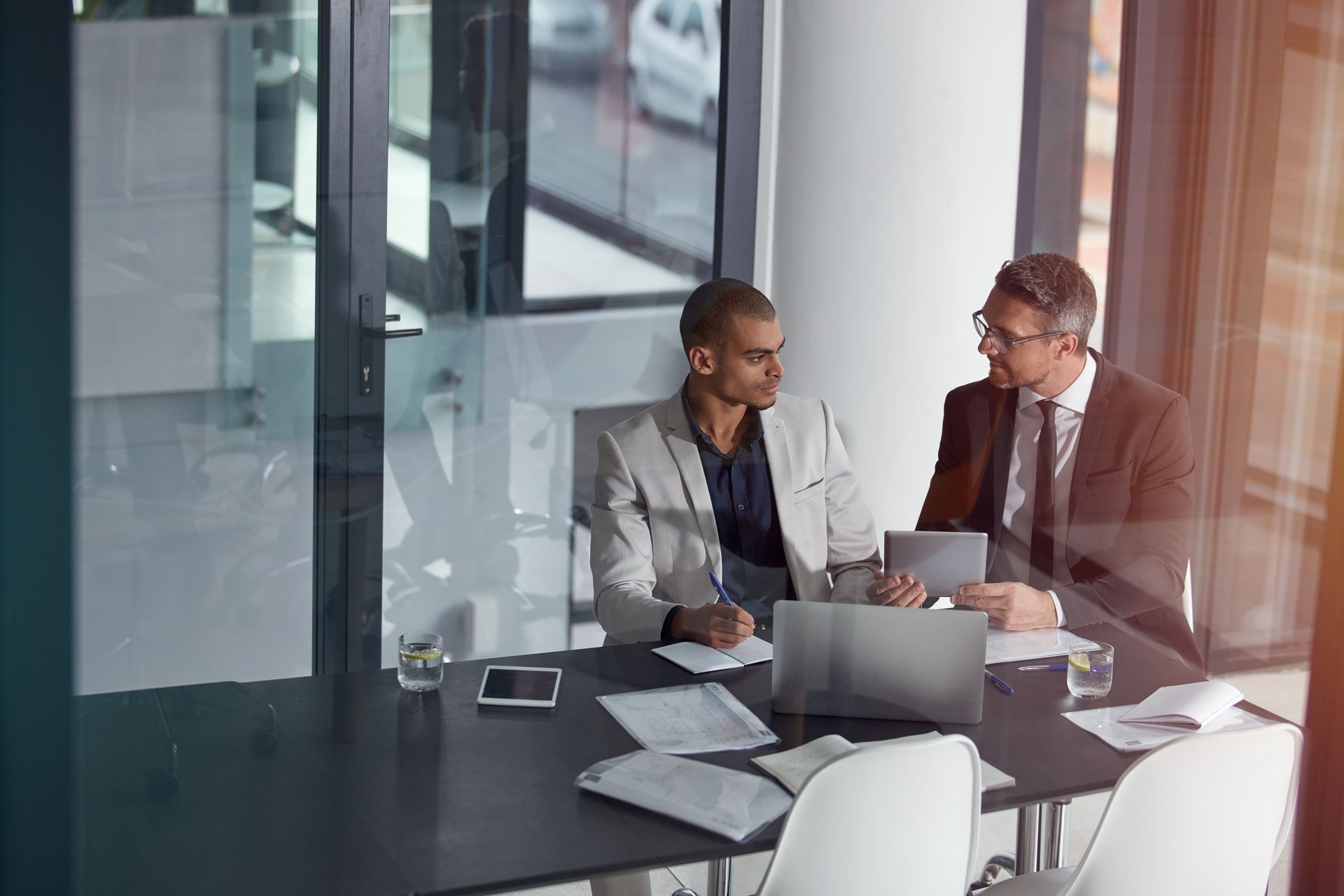 Two coworkers in a glass office meeting, discussing documents with a laptop on the table.