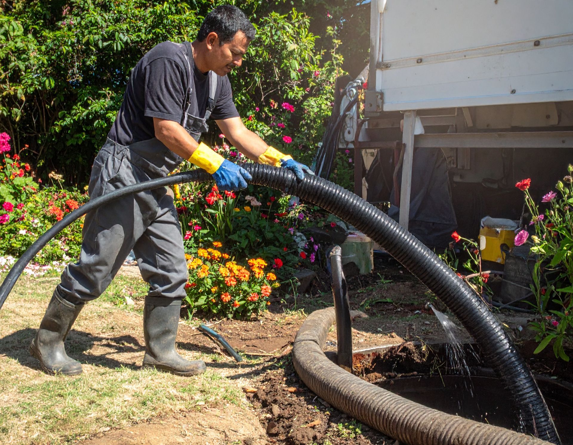 Person in gloves uses a hose to clean a yard beside flowers and a white truck.