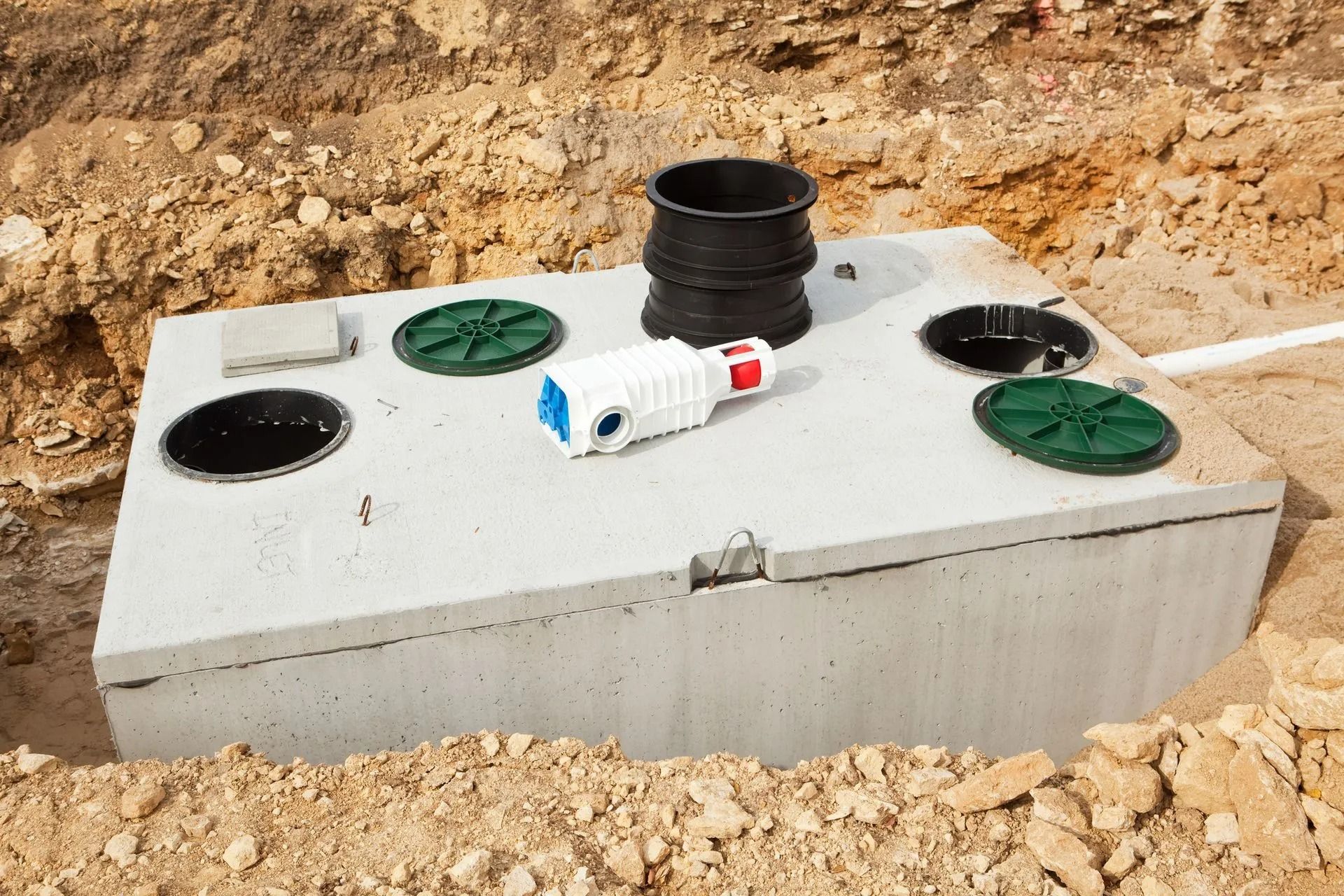 A concrete septic tank set in an excavated dirt trench, featuring black risers, green access covers, and a white pump part.