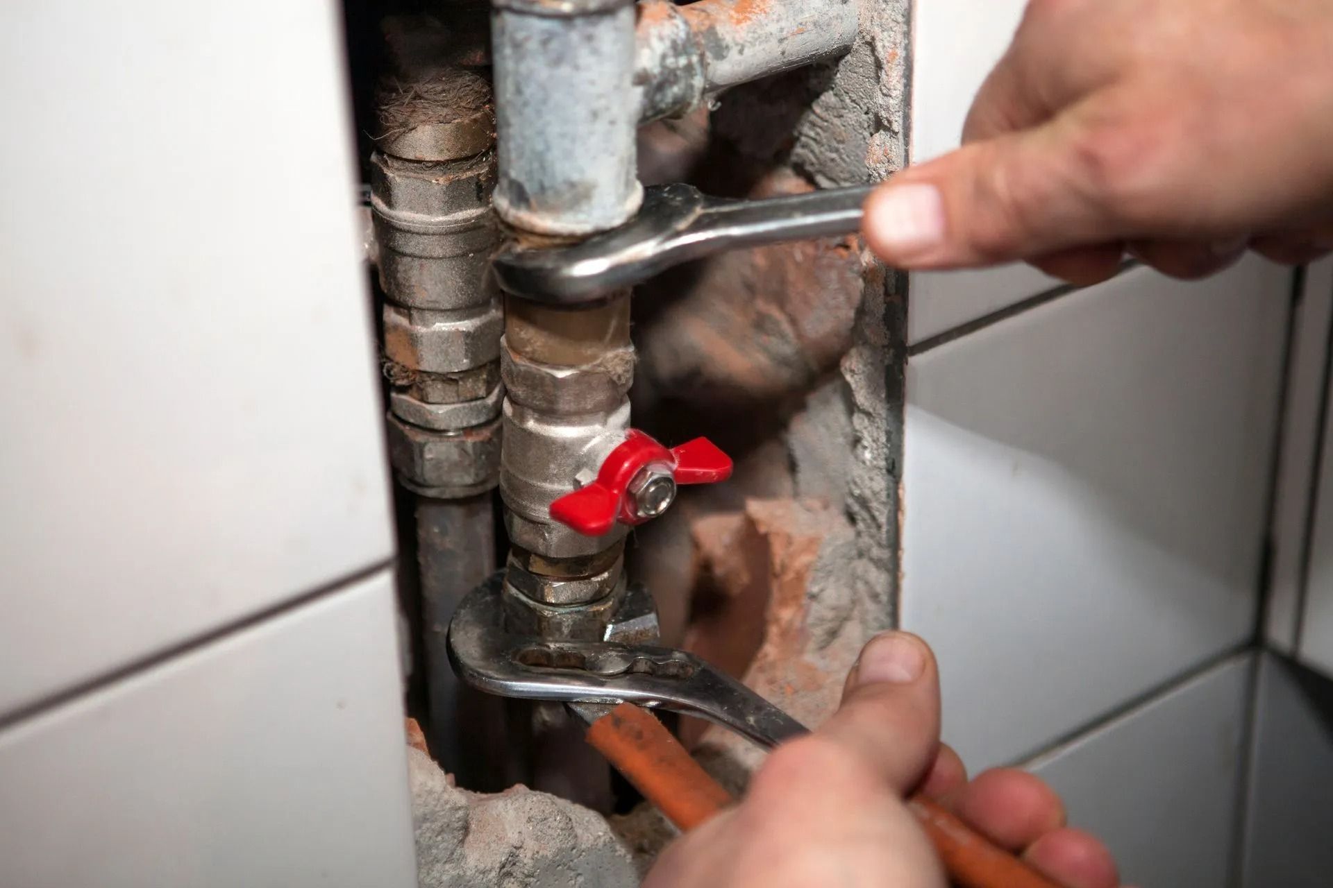 Two hands use wrenches to tighten a metal pipe fitting with a red valve handle inside a tiled wall recess.