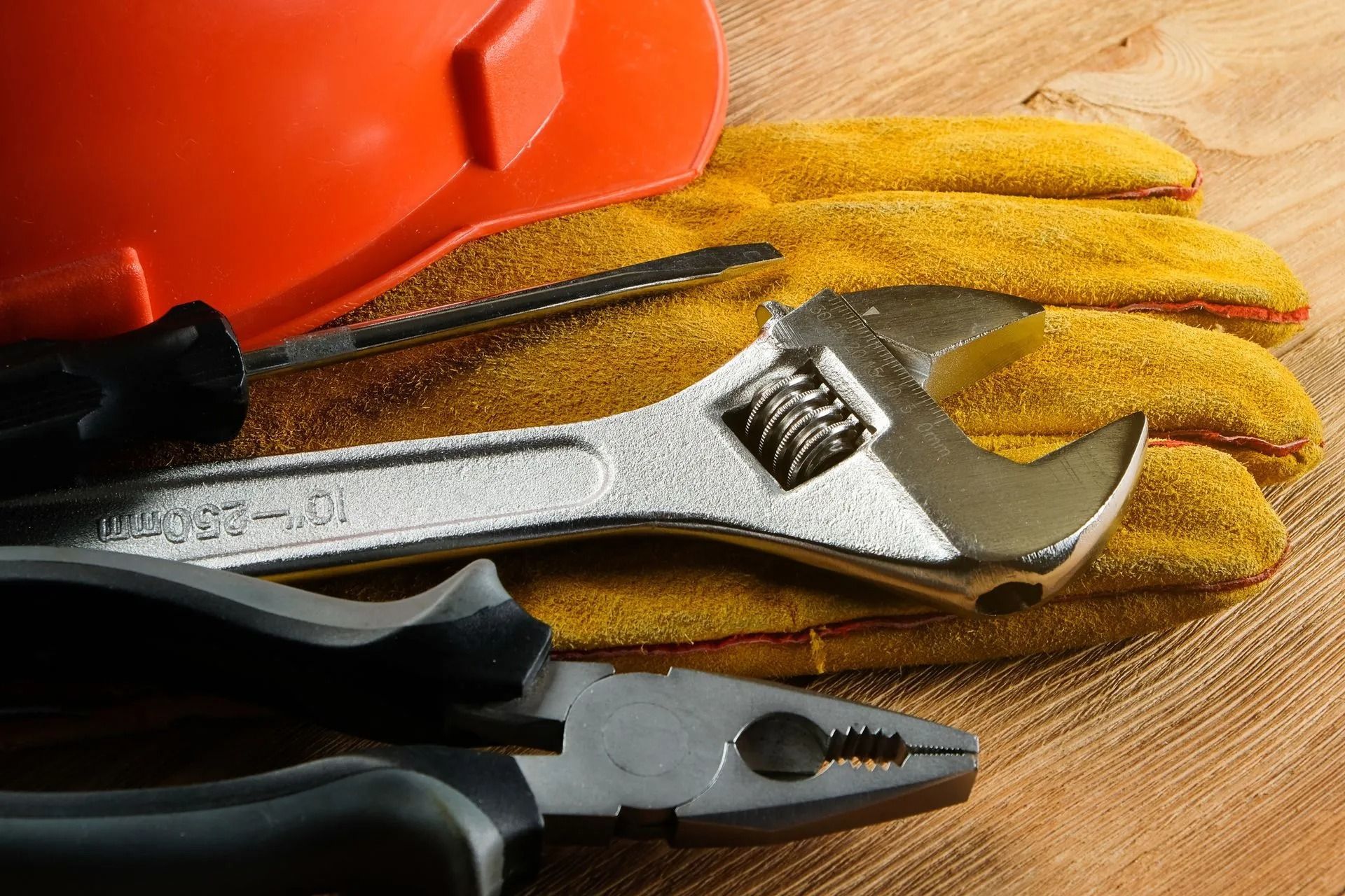 An orange hard hat, leather gloves, an adjustable wrench, pliers, and a screwdriver on a wooden surface.