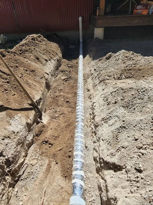 A trench in the ground containing a long, silver, flexible duct pipe running from a building structure.