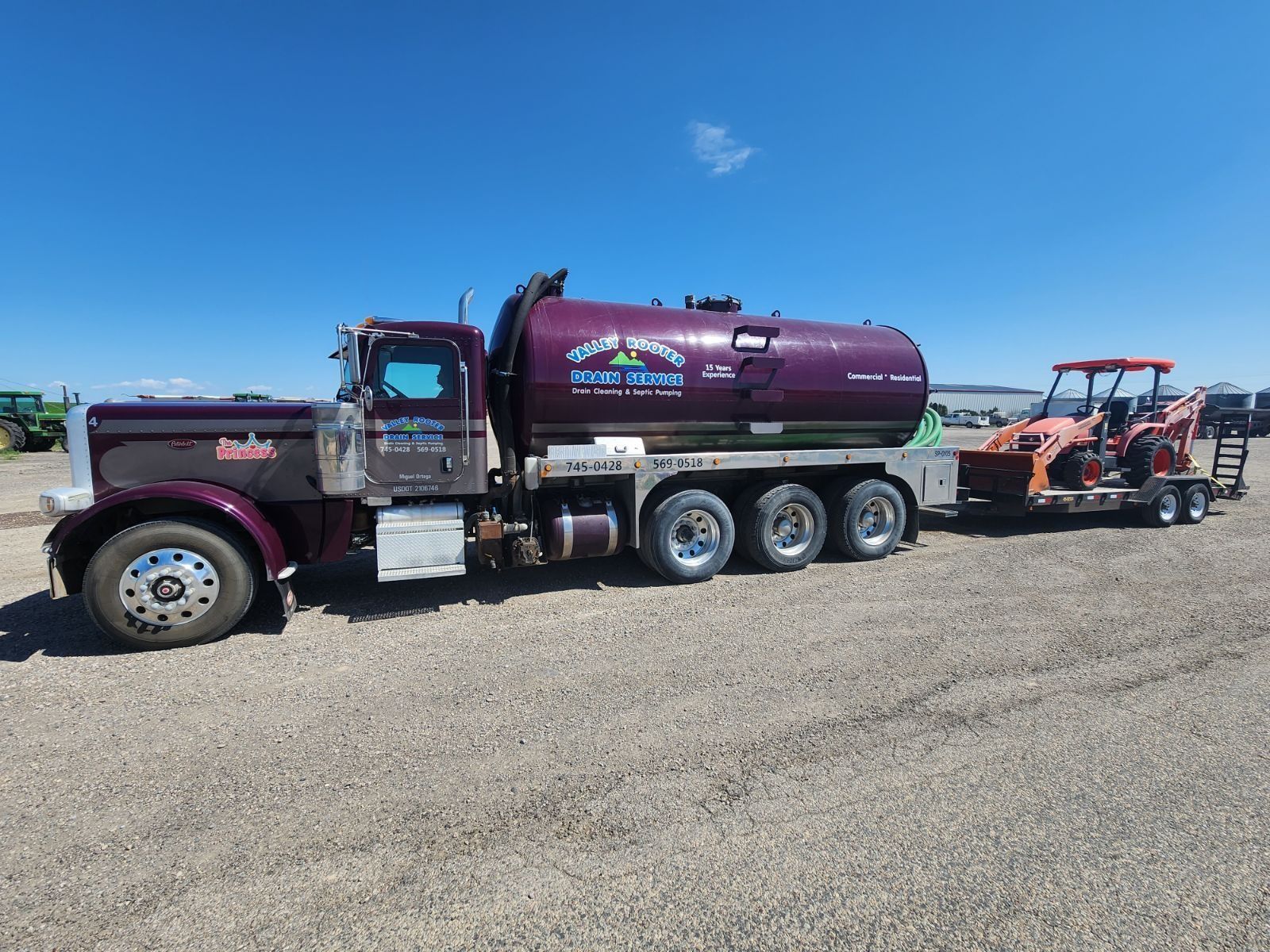 A purple tanker truck parked on a gravel lot, towing a trailer with an orange piece of construction equipment.