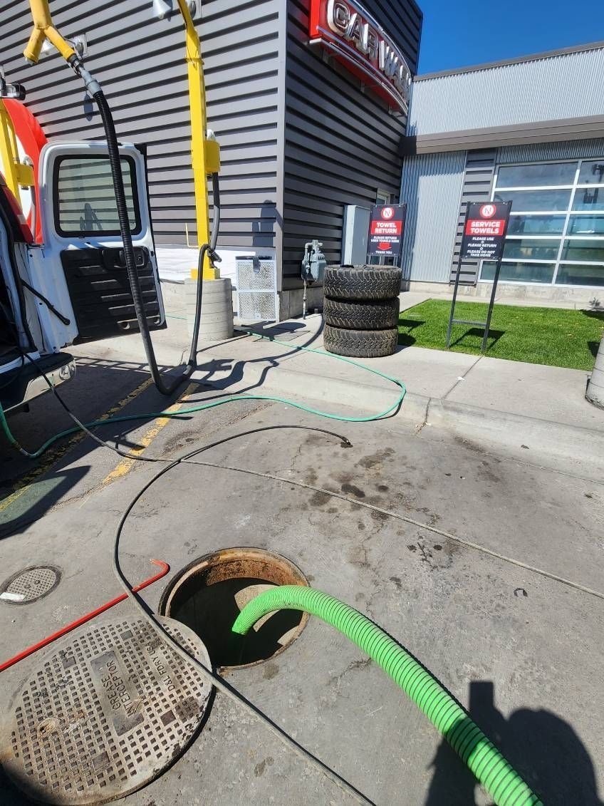 A bright green suction hose connected to a service truck sits inside an open manhole in a parking lot near a building.