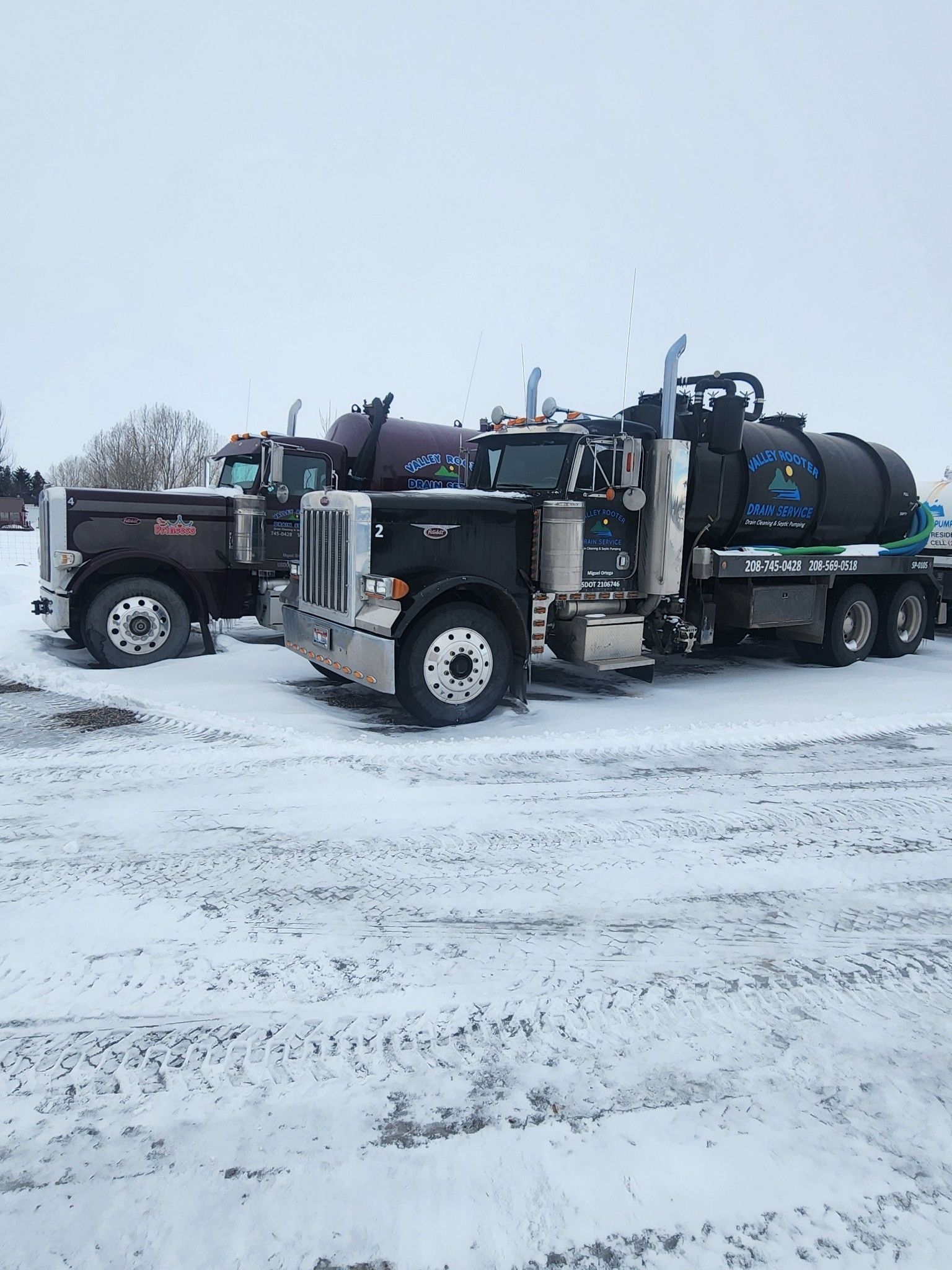Two black industrial vacuum trucks parked on a snow-covered gravel lot.