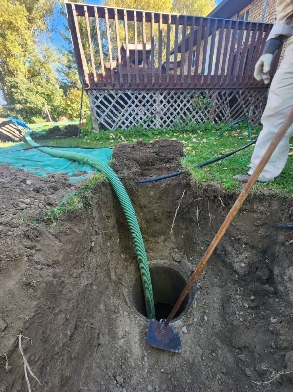 A person holding a shovel beside a hole in the ground where a green suction hose is being used to pump out a septic tank.