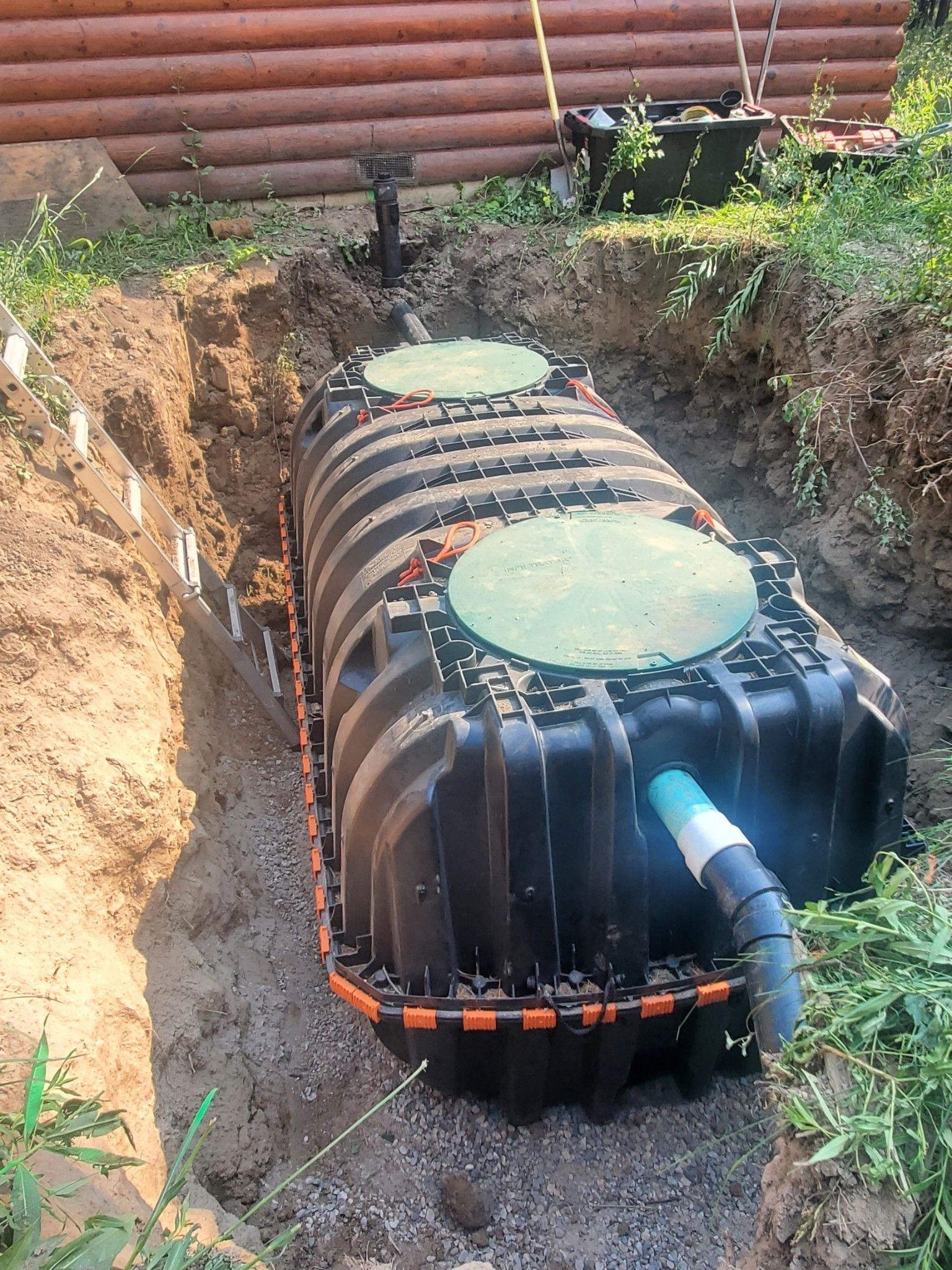 A black, ribbed plastic septic tank installed in a dirt excavation trench near a wooden building, with green access lids.