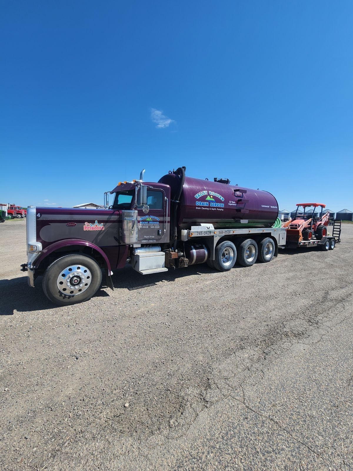 A burgundy vacuum tank truck parked on a gravel lot under a clear blue sky, hauling a trailer with construction equipment.