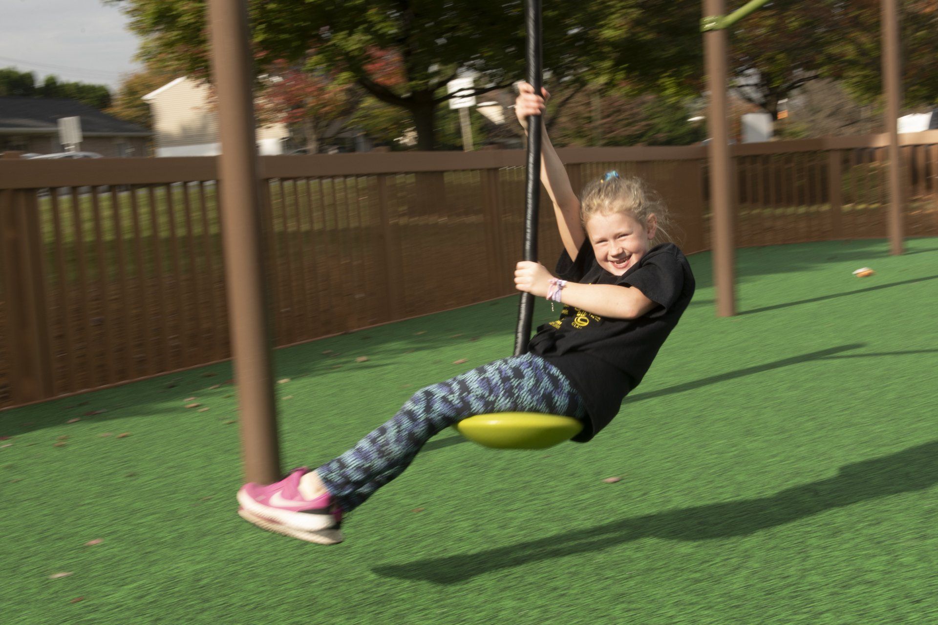 Girl riding a zip line at a playground, smiling, wearing black shirt, colorful pants, pink shoes, green surface.