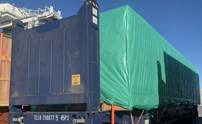Blue shipping container with a green tarp-covered rectangular object on top, outdoors, under a clear sky.