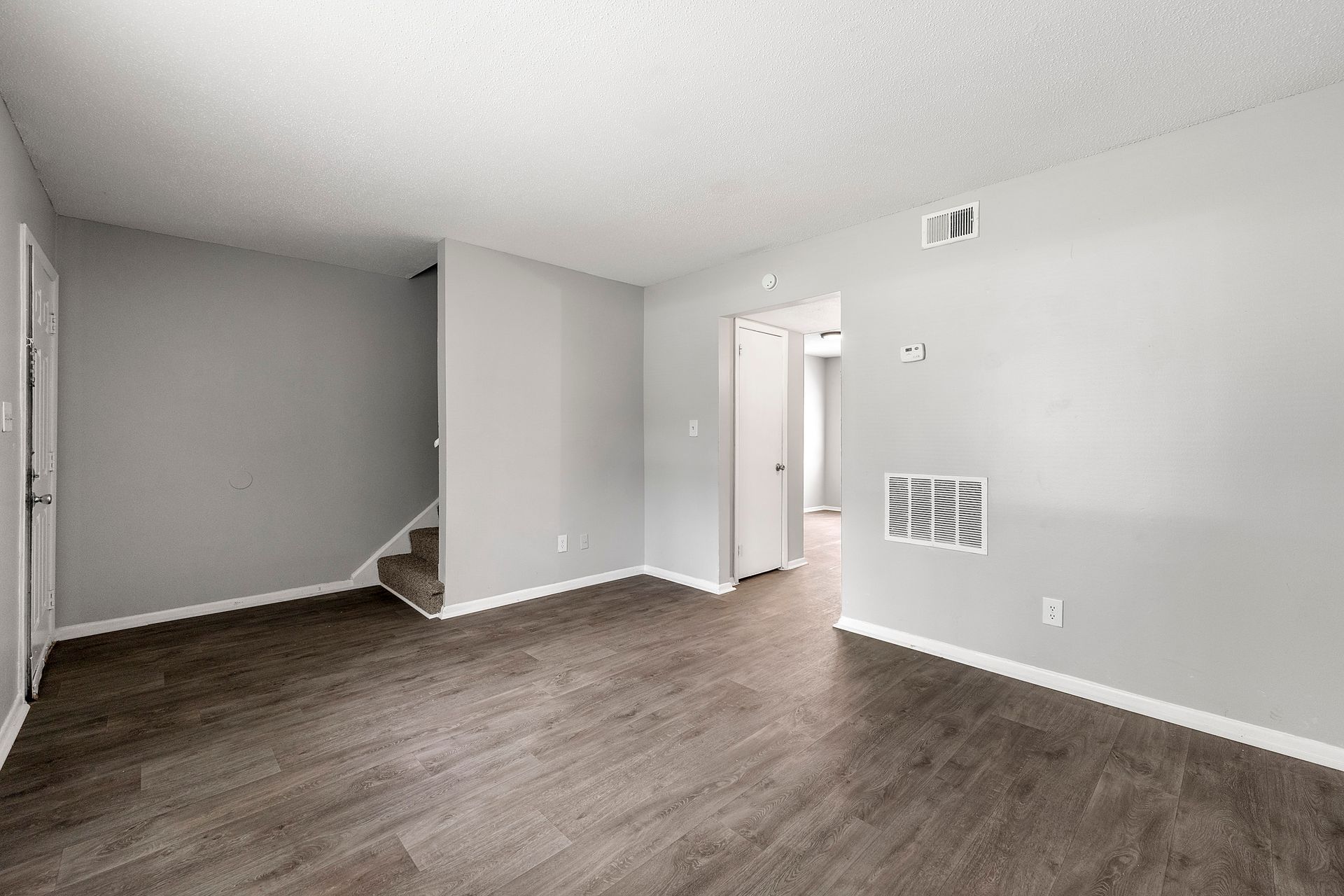 Empty gray living room with wood floors, white trim, and a staircase in the corner