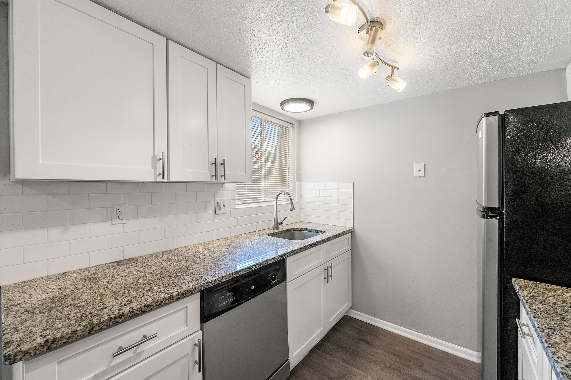 Bright galley kitchen with white cabinets, granite counters, stainless dishwasher, and black refrigerator