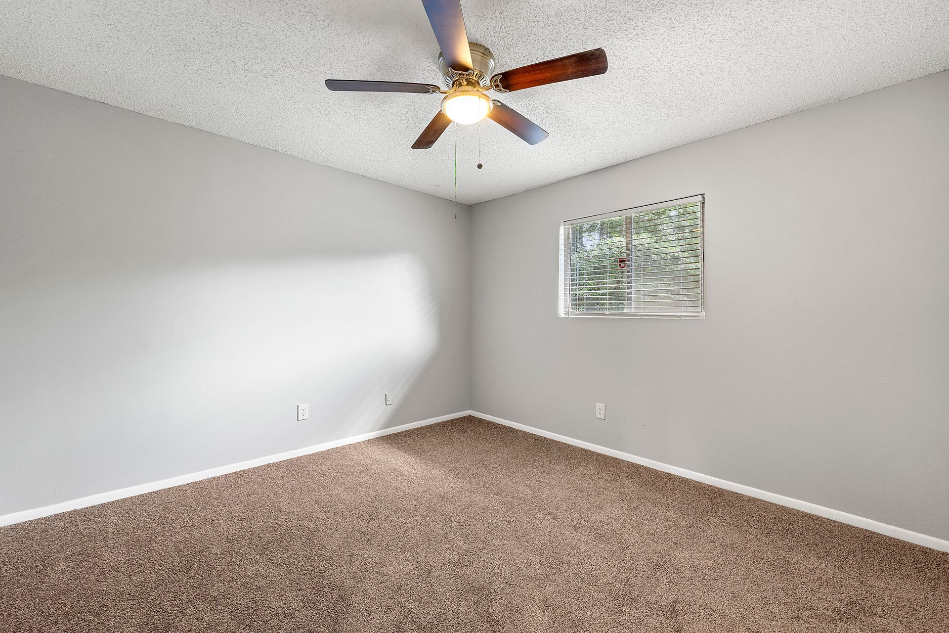 Empty beige-carpeted bedroom with gray walls, a ceiling fan, and a small window.