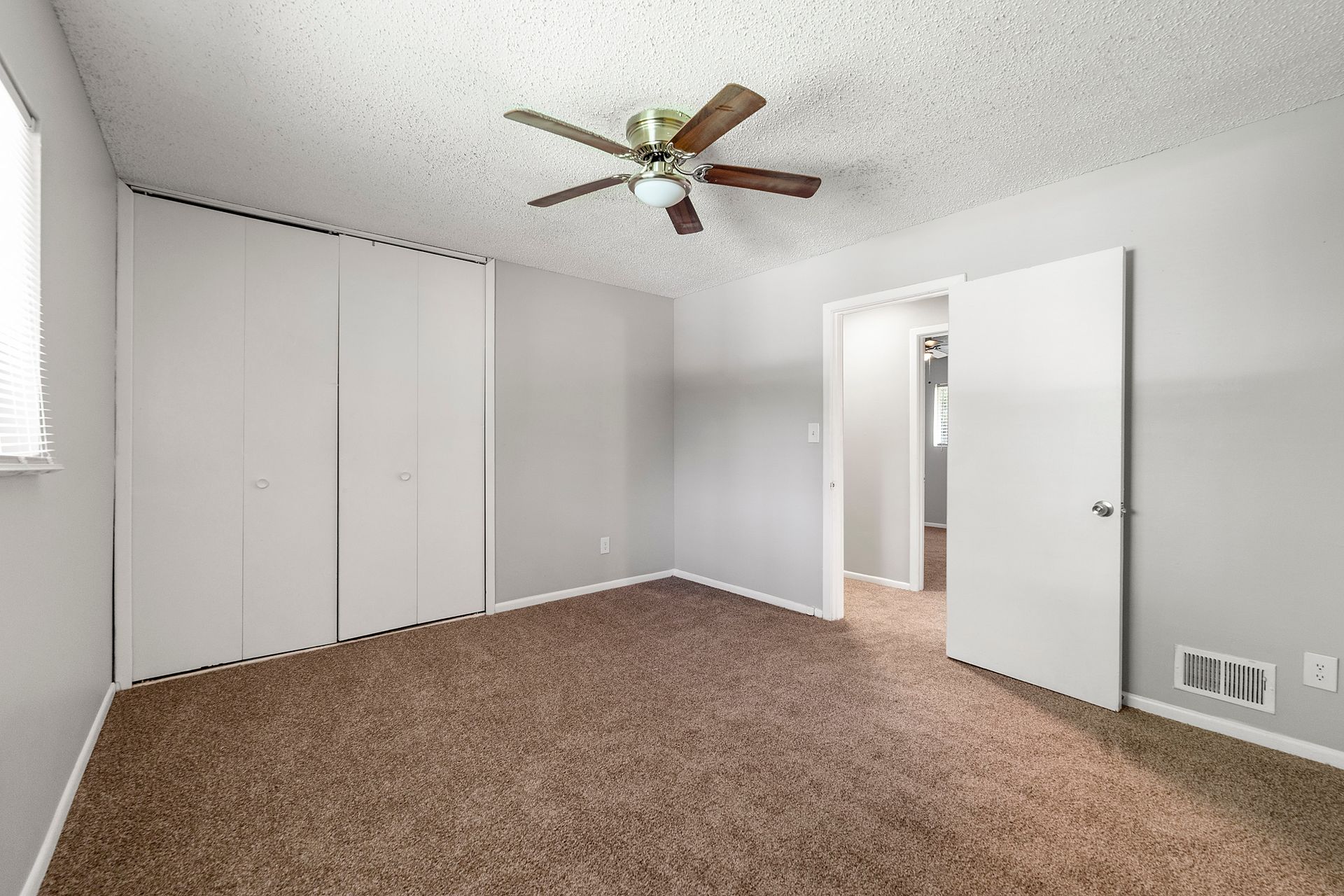 Empty carpeted bedroom with white walls, closet doors, a ceiling fan, and an open door to a hallway
