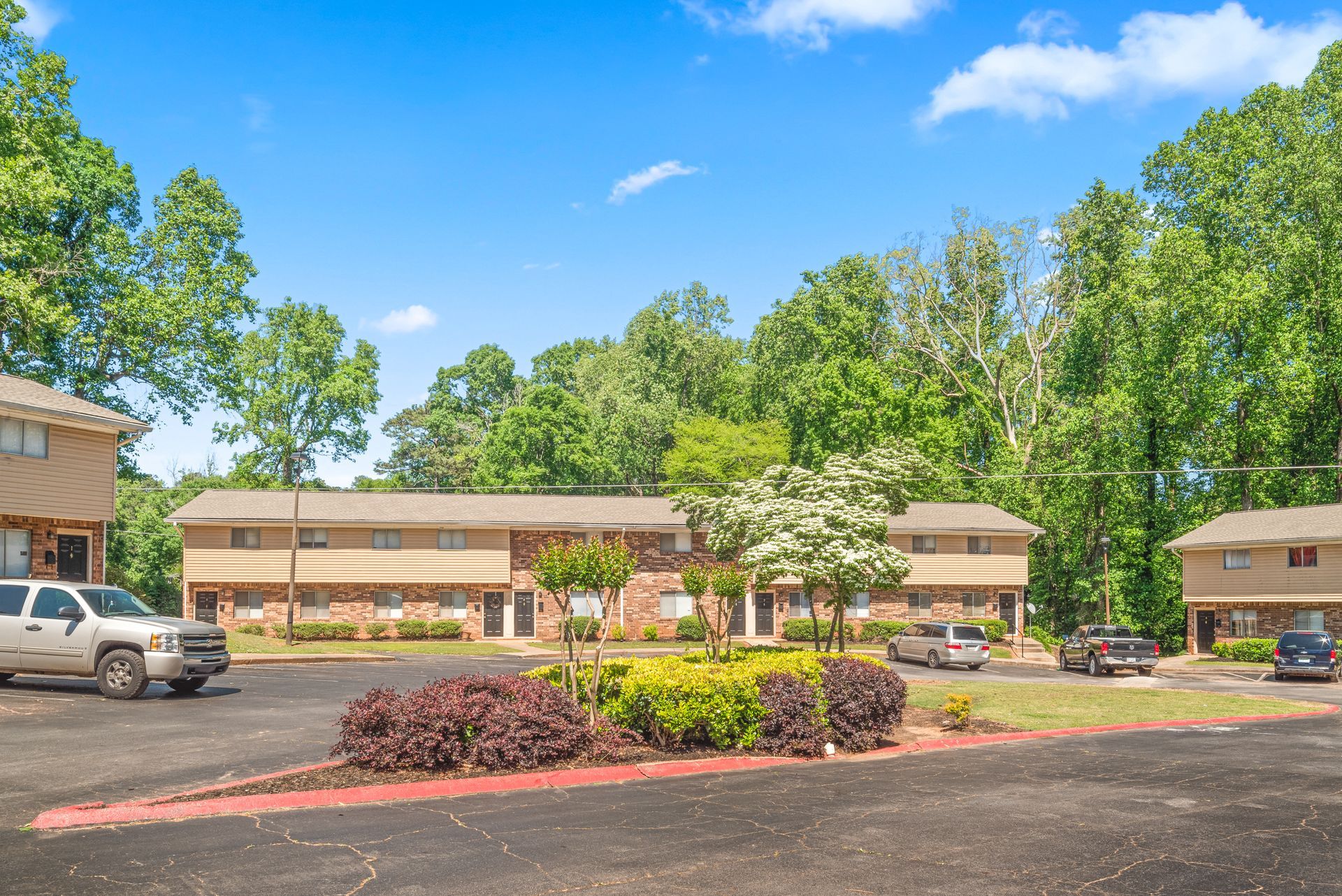 Two-story apartment building with parked cars, trees, and a bright blue sky
