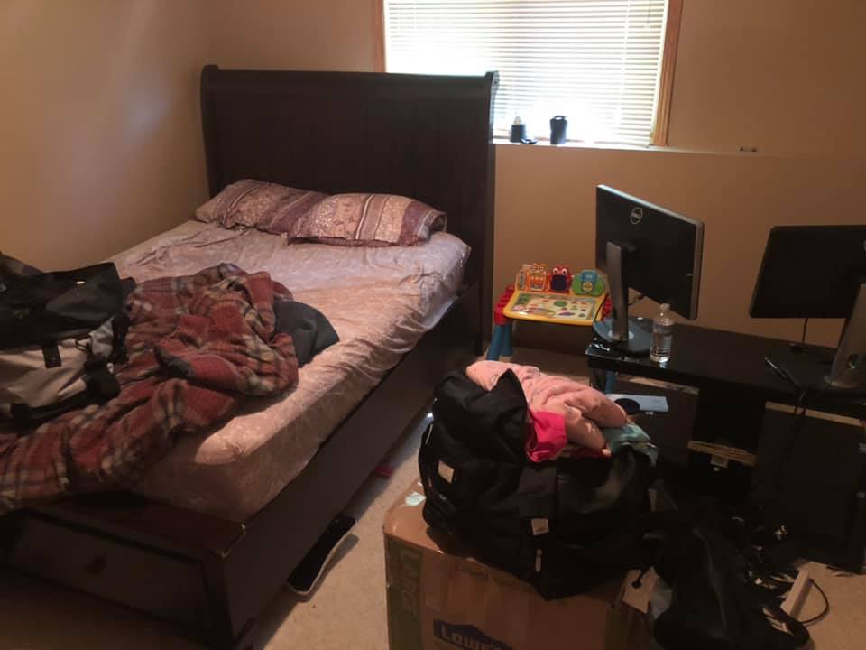 A bedroom with a dark wooden bed, a desk featuring two computer monitors, and a cardboard box topped with a black bag.