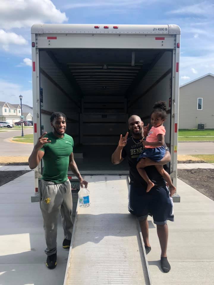 Two people stand at the back of an open moving truck in a residential driveway, with one holding a child.