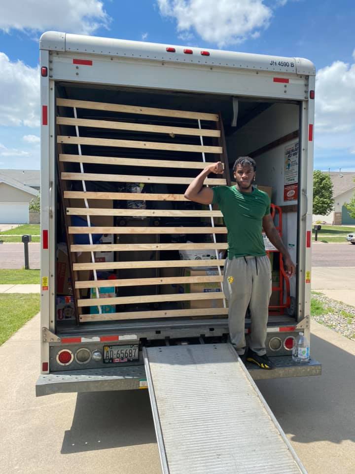 A person in a green shirt and gray sweatpants flexes their arm in front of a wooden bed frame inside a moving truck.
