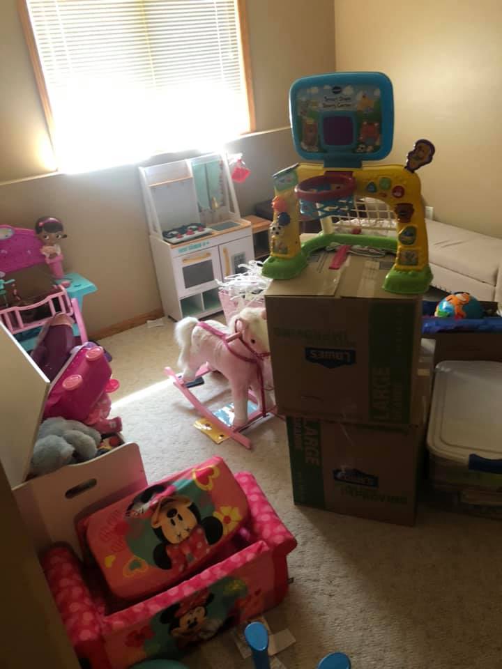 A child's playroom featuring a toy basketball hoop on stacked cardboard boxes, a rocking horse, and a Minnie Mouse sofa.