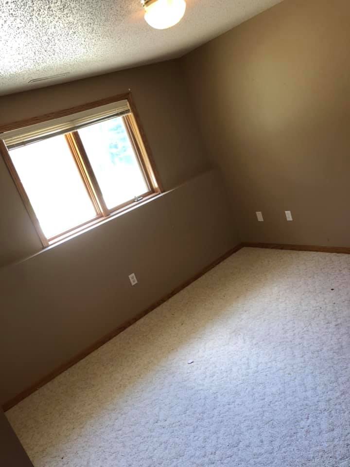 An empty room with tan walls, light-colored carpet, and a window with blinds under a ceiling light.
