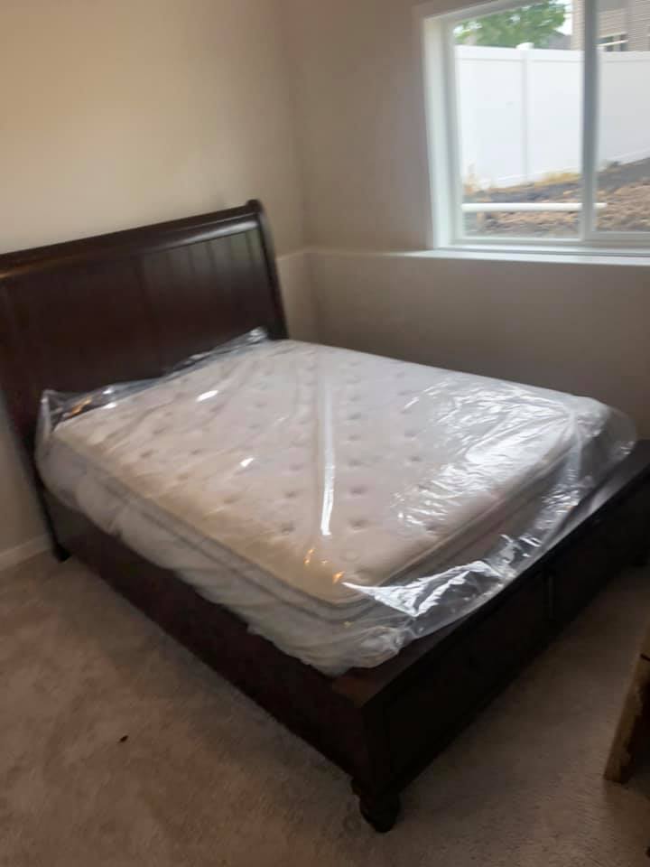 A dark wood bed frame with a mattress still covered in protective plastic, situated in a carpeted room by a window.