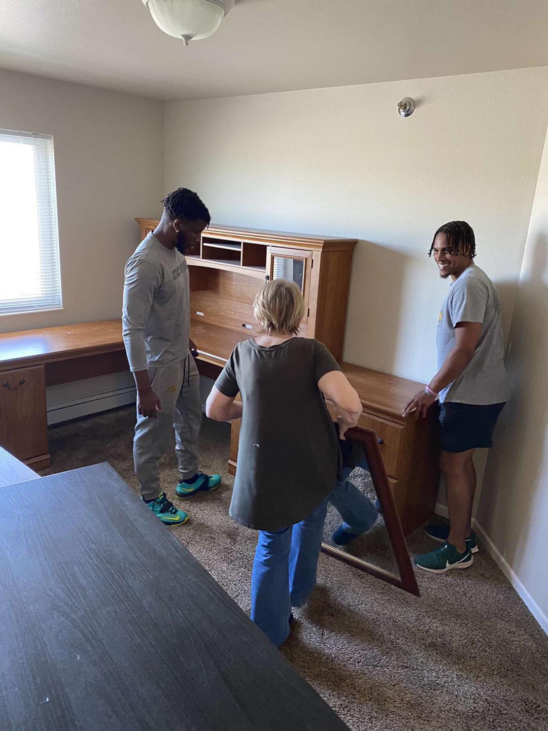 Three people move a large wooden desk in a room with beige walls, a window, and brown carpet.
