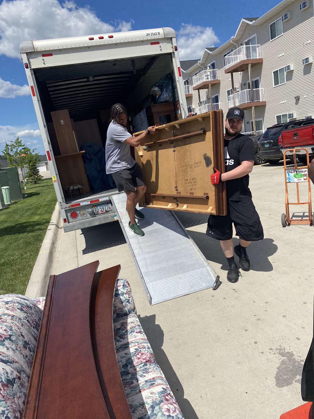 Two people moving a large wooden tabletop from the back of a rental truck down a metal ramp on a sunny day.