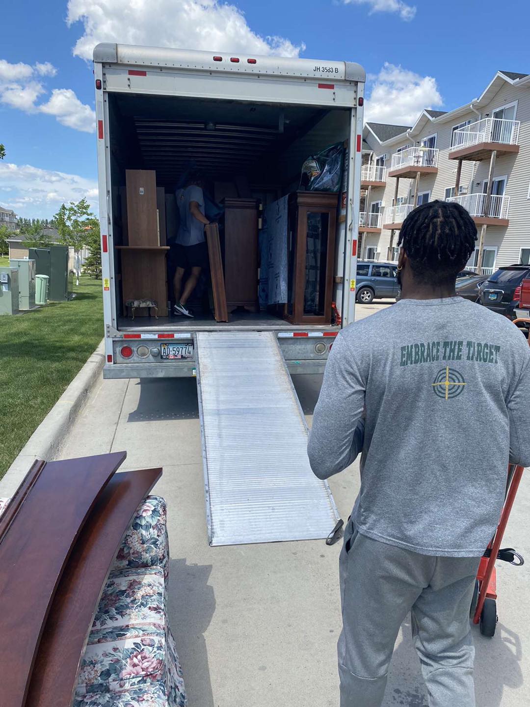 A person moves furniture into a parked moving truck using a ramp on a sunny day near an apartment building.