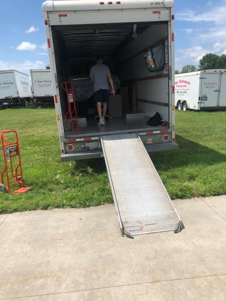 A person in a gray shirt and shorts walks into the back of a moving truck with a deployed ramp on a grassy lot.