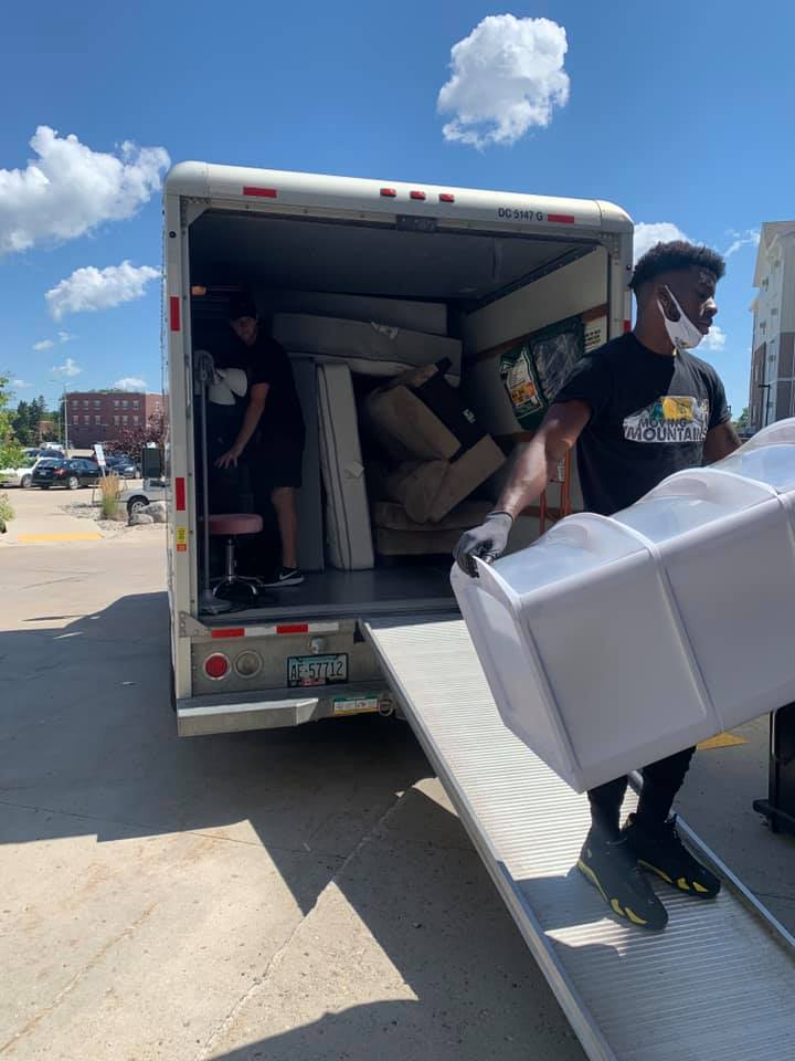 Two movers load furniture into a box truck on a sunny day. One carries a large white box down a ramp.
