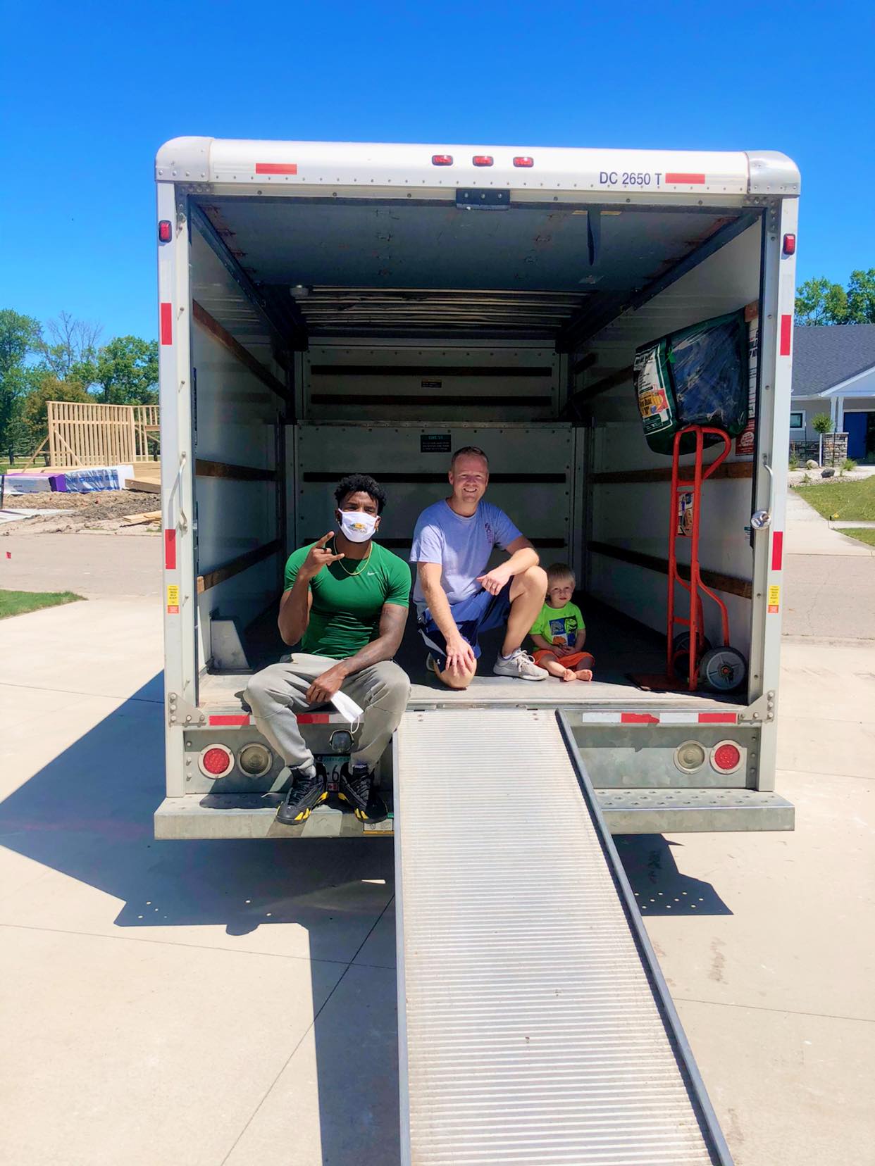 Two people sit inside the back of an open moving truck with a ramp on a sunny day near a residential area.