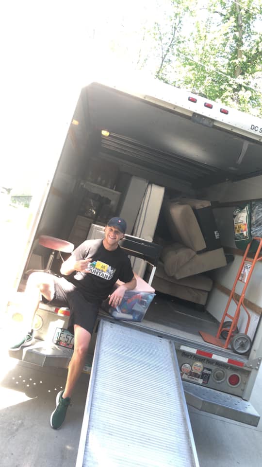 A person sits on a moving truck ramp, smiling, with a loaded truck interior and dolly in the background.