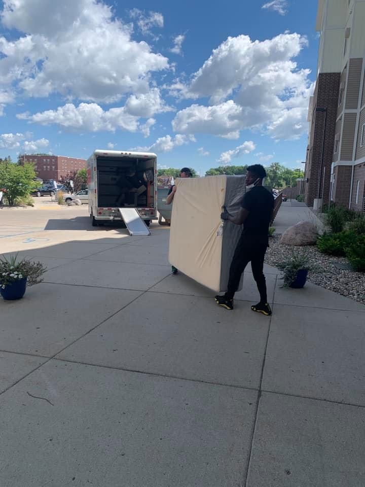 Two people carry a large mattress on a paved walkway toward a moving truck parked on a sunny day.