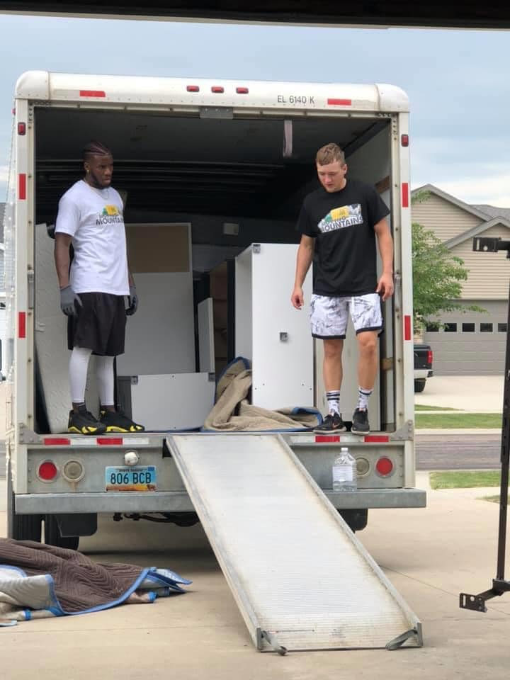 Two people in athletic gear stand inside the back of a moving truck with a ramp extended to the driveway.