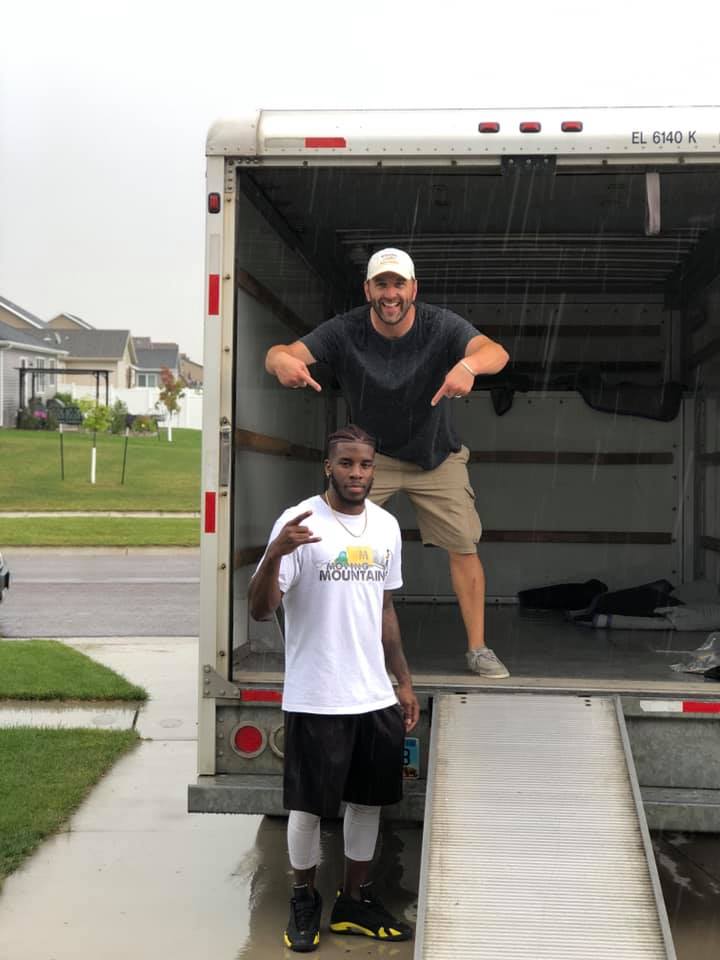 Two people stand at the back of an open moving truck; one stands inside pointing down, the other stands outside posing.