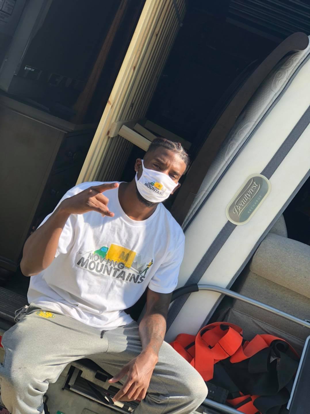 A person wears a white t-shirt with a mountain graphic, a face mask, and grey sweatpants, sitting near a white mattress.