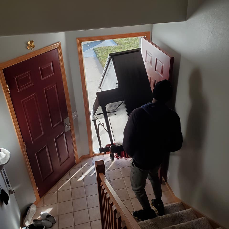 A person in dark clothing stands on indoor stairs, holding the door open for a large dark piece of furniture being moved in.