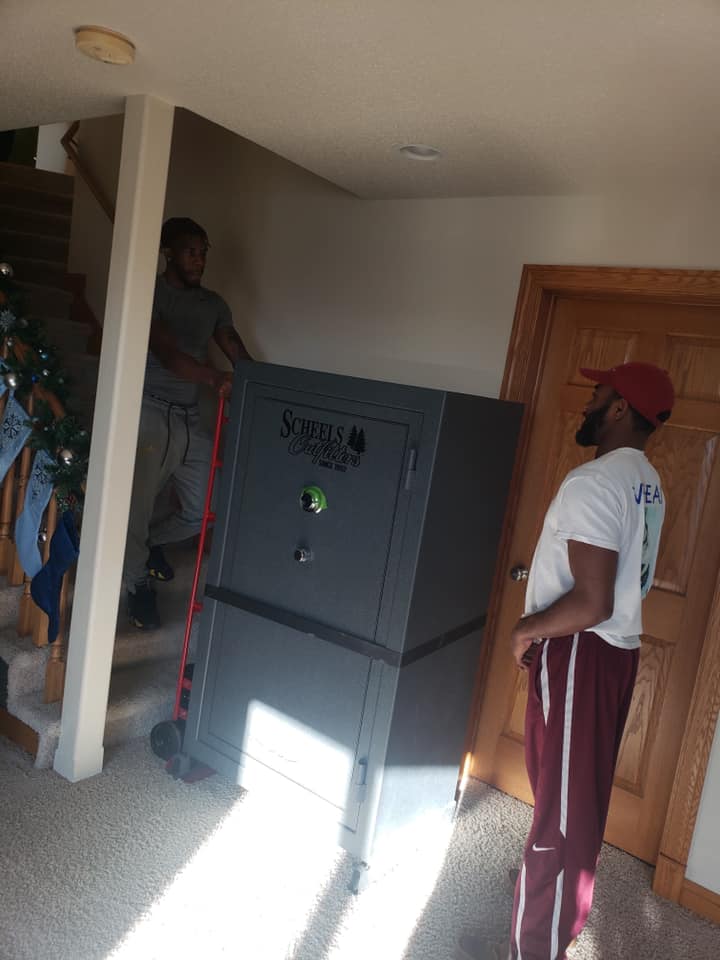 Two people move a large, dark gray SureLock Security safe into a home near a wooden door and a staircase.
