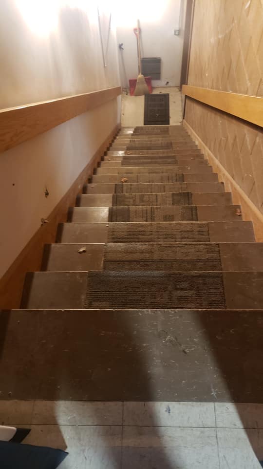 A steep, narrow wooden staircase with brown carpet runners, leading down to a landing with a broom and a light switch.